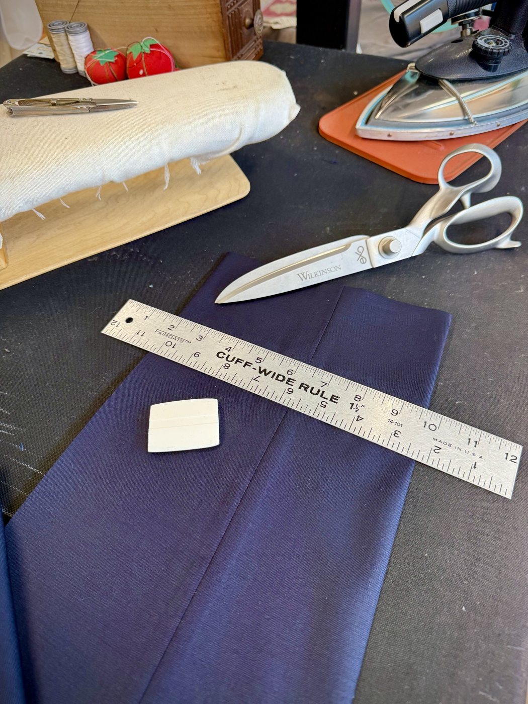 Tailor shears, a steel cuff-wide ruler and tailor chalk resting on navy trouser fabric at the pressing table